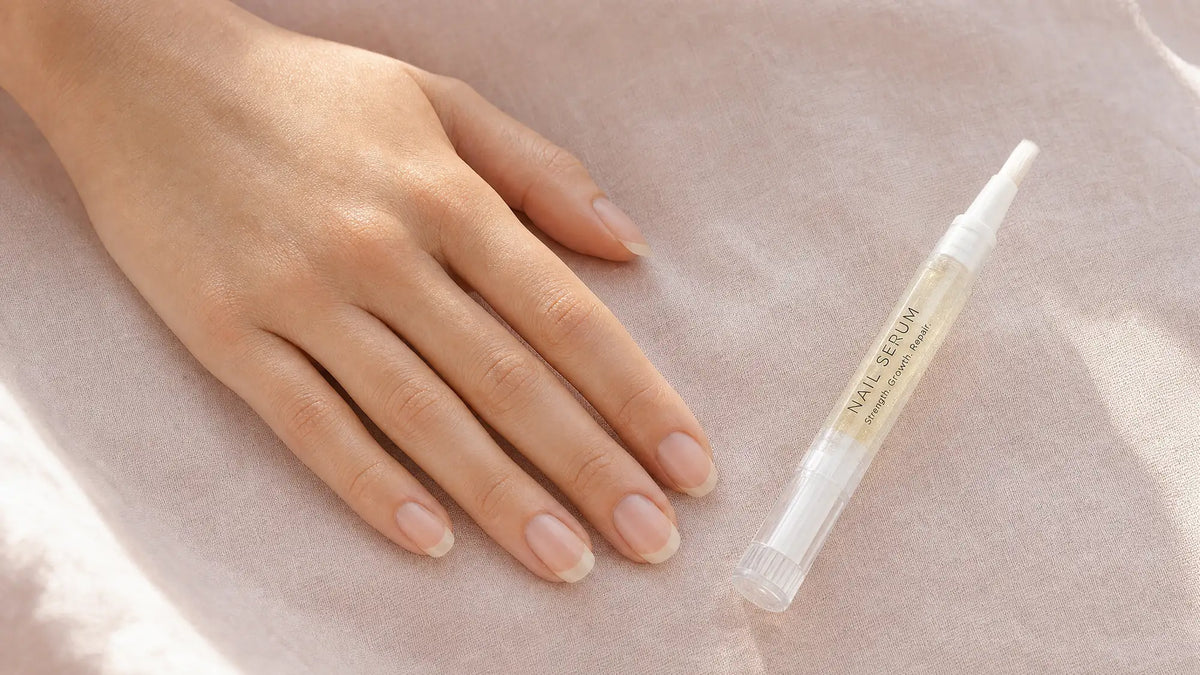 Long healthy natural nails resting on blush linen beside a Body Collective nail serum pen