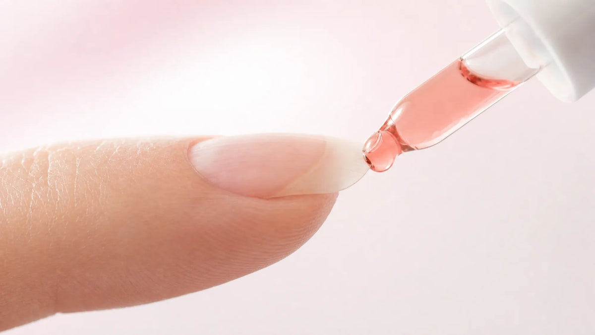 A natural nail seen from the side with the pink nail bed showing through the plate, and a dropper placing a drop of serum at the free edge