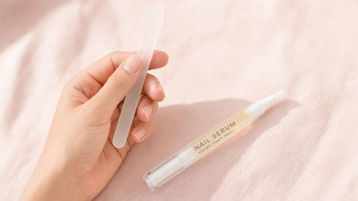Short smooth natural nails holding a glass file beside a Body Collective pen