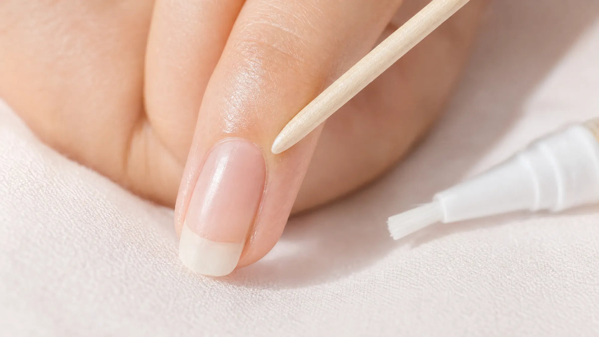 Close up of an orangewood stick resting at the base of a fingernail, with the brush of a nail serum pen beside it